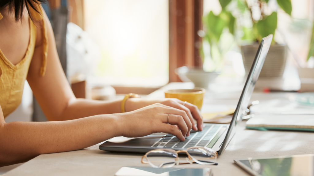 01 Create a shop A woman in a yellow top types on a laptop in a sunlit room. A potted plant, glasses, and a smartphone are on the desk.