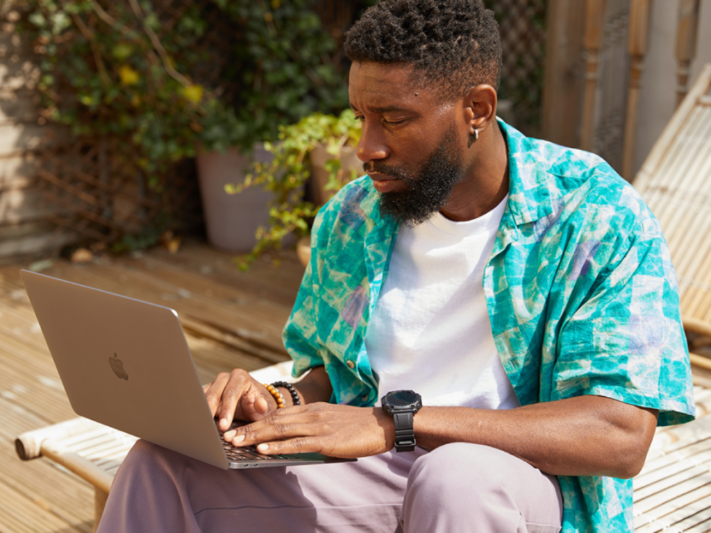 01 How to choose a profitable t shirt niche Man in a colorful shirt focuses on a laptop while sitting on a sunlit deck. He appears thoughtful.