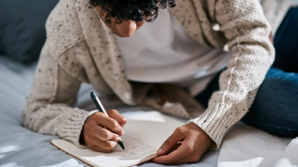 02 Name your Etsy store A man with curly hair is lying on a bed, wearing a cozy sweater, focused on writing in a notebook.