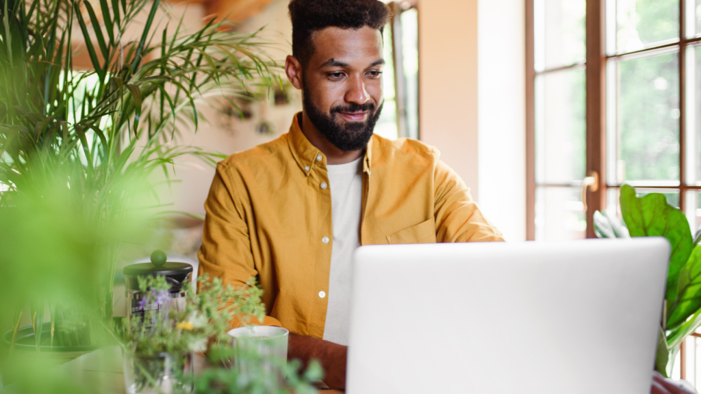 04 Review customize and launch your store A man in a yellow shirt works on a laptop surrounded by lush green plants, near a sunlit window.