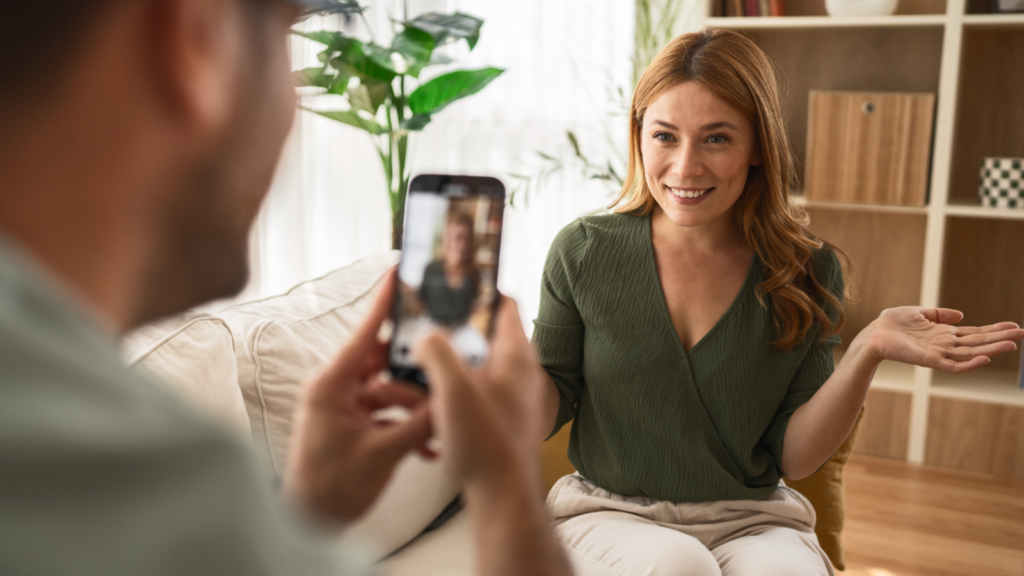 05 Build visibility with social media A woman with red hair smiles warmly, sitting on a couch indoors, while a person films her using a smartphone.