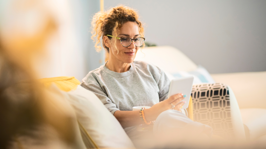 06 Tips on how to start a business on Etsy successfully A woman with curly hair and glasses sits on a couch, relaxed, reading a tablet. She wears a gray sweater.