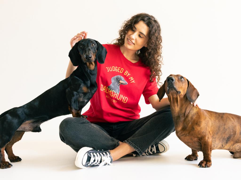 07 Pet and animal lovers A woman in a red shirt with a dachshund graphic sits cross-legged, smiling at two dachshunds.
