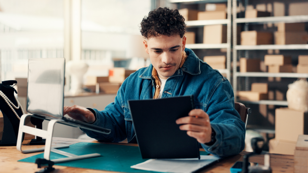07 Stay active and publish regularly A man in a denim jacket is working at a desk with a tablet and a laptop in a warehouse. Shelves with boxes are visible in the background.