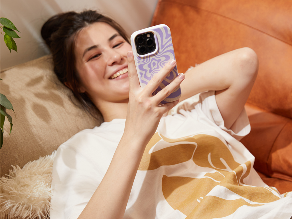 12 Sell custom t shirts with Printify A woman lies on a couch, smiling at her phone with a purple-patterned case. She's wearing a cream shirt with a gold print.