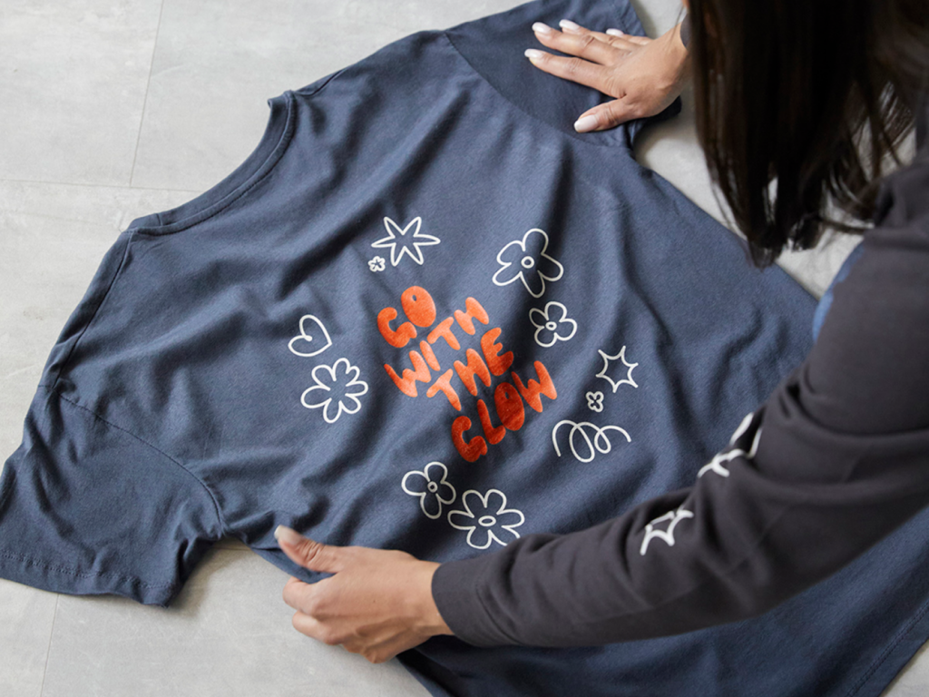 16 Streetwear and fashion A person arranges a navy t-shirt with the phrase "Go With the Glow" in orange, surrounded by white doodle flowers and stars.