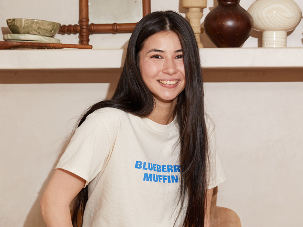 18 Put your favorite font on a custom t shirt with Printify A woman with long dark hair is smiling, wearing a cream "Blueberry Muffin" t-shirt. Behind are decorative ceramics on a wooden shelf.