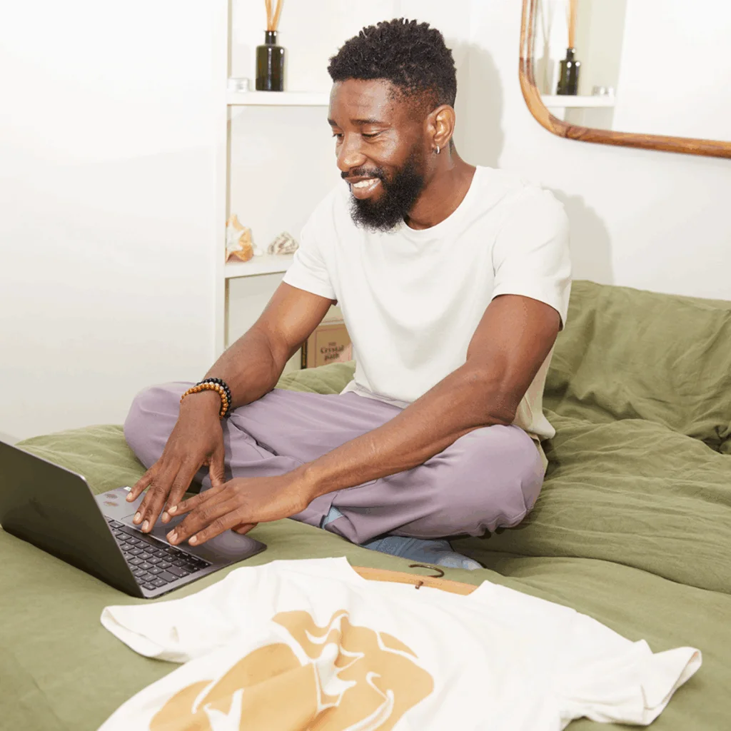 A man sitting cross legged on a bed with a t shirt beside him using a laptop to find the best shirts for sublimation printing A man sitting cross-legged on a bed, with a t-shirt beside him, using a laptop to find the best shirts for sublimation printing