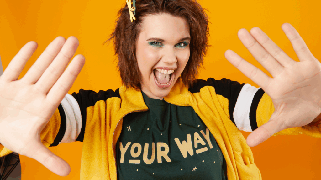 Decide what to sell A woman with green eyeshadow smiling broadly, wearing a yellow and black jacket and a "Your Way" t-shirt.