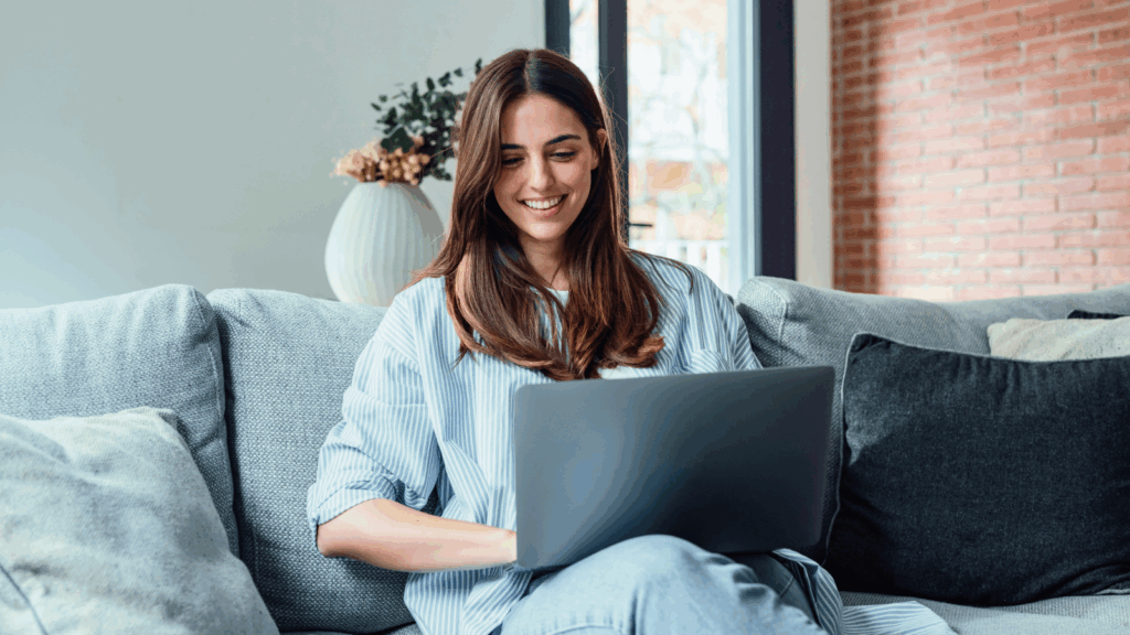 Decide where to sell and build your store How to start a clothing brand A smiling woman is sitting on a couch with a laptop on her lap.