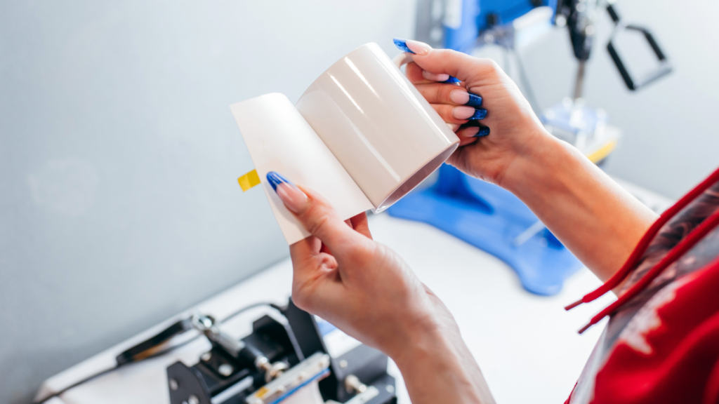 Essential equipment for the sublimation process A person with blue-painted nails holds a plain white mug and a piece of paper, likely for customization, in a workshop with equipment in the background.