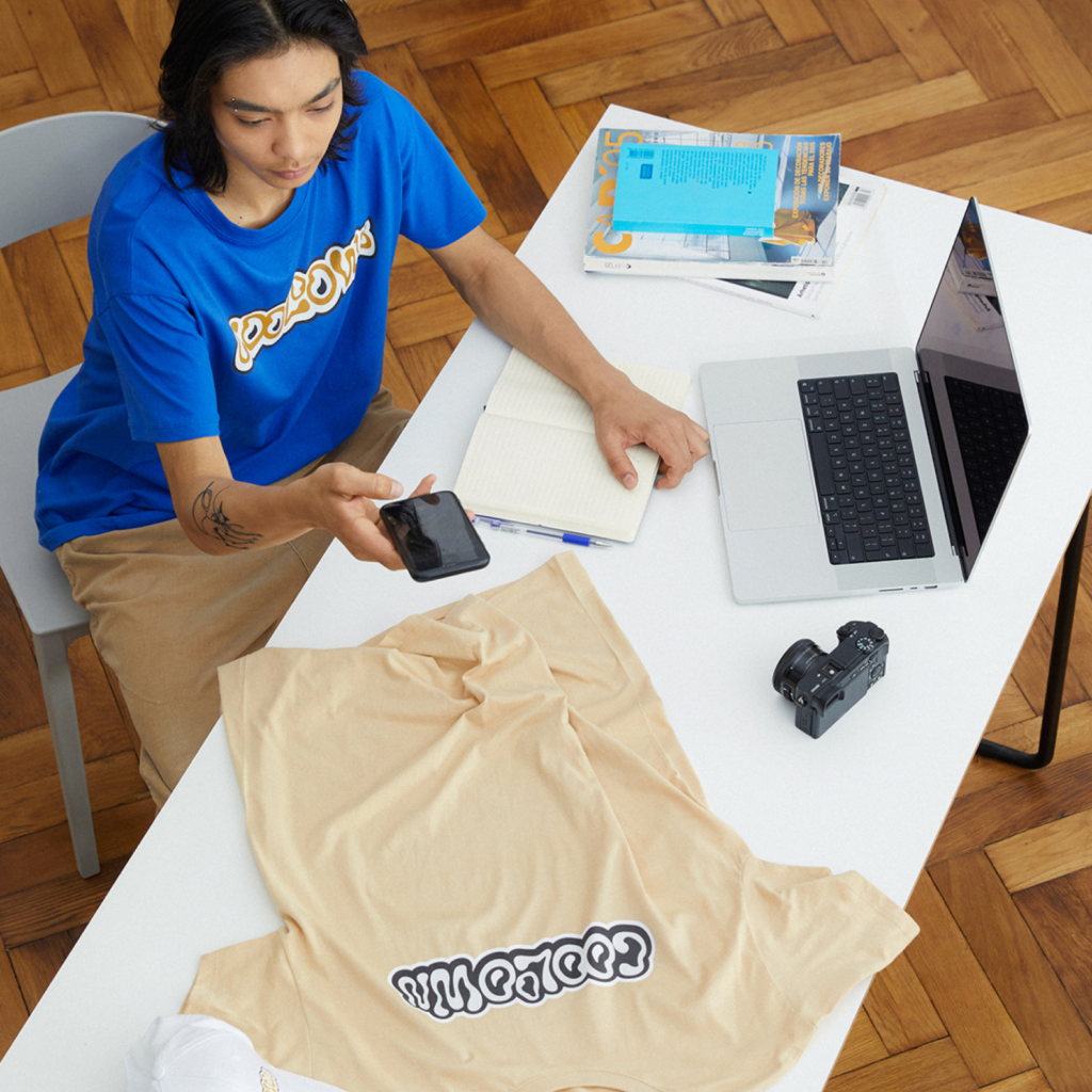 How does Printify Pop Up Store work with Print on Demand A man in a blue t-shirt sits at a white desk. He’s taking a picture of a beige t-shirt, surrounded by a notebook, camera, and open laptop.