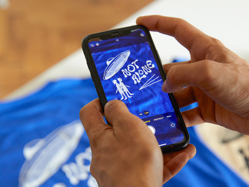 Take beautiful product images A person taking a picture of a custom blue “Not Alone” t-shirt using a mobile phone.