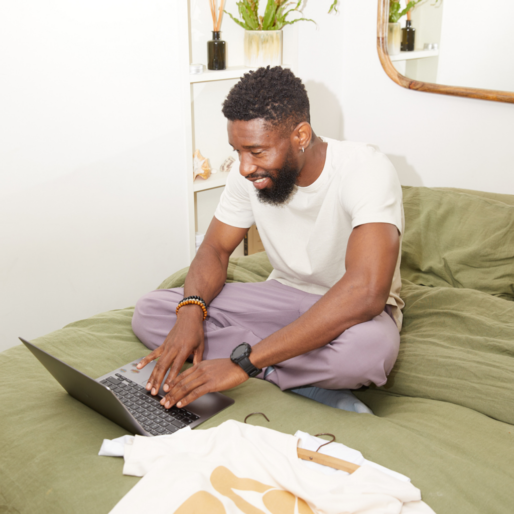 What is Printify Pop Up Store Man sitting cross-legged on a bed with green sheets, smiling while using a laptop. He's wearing a white t-shirt and purple pants.